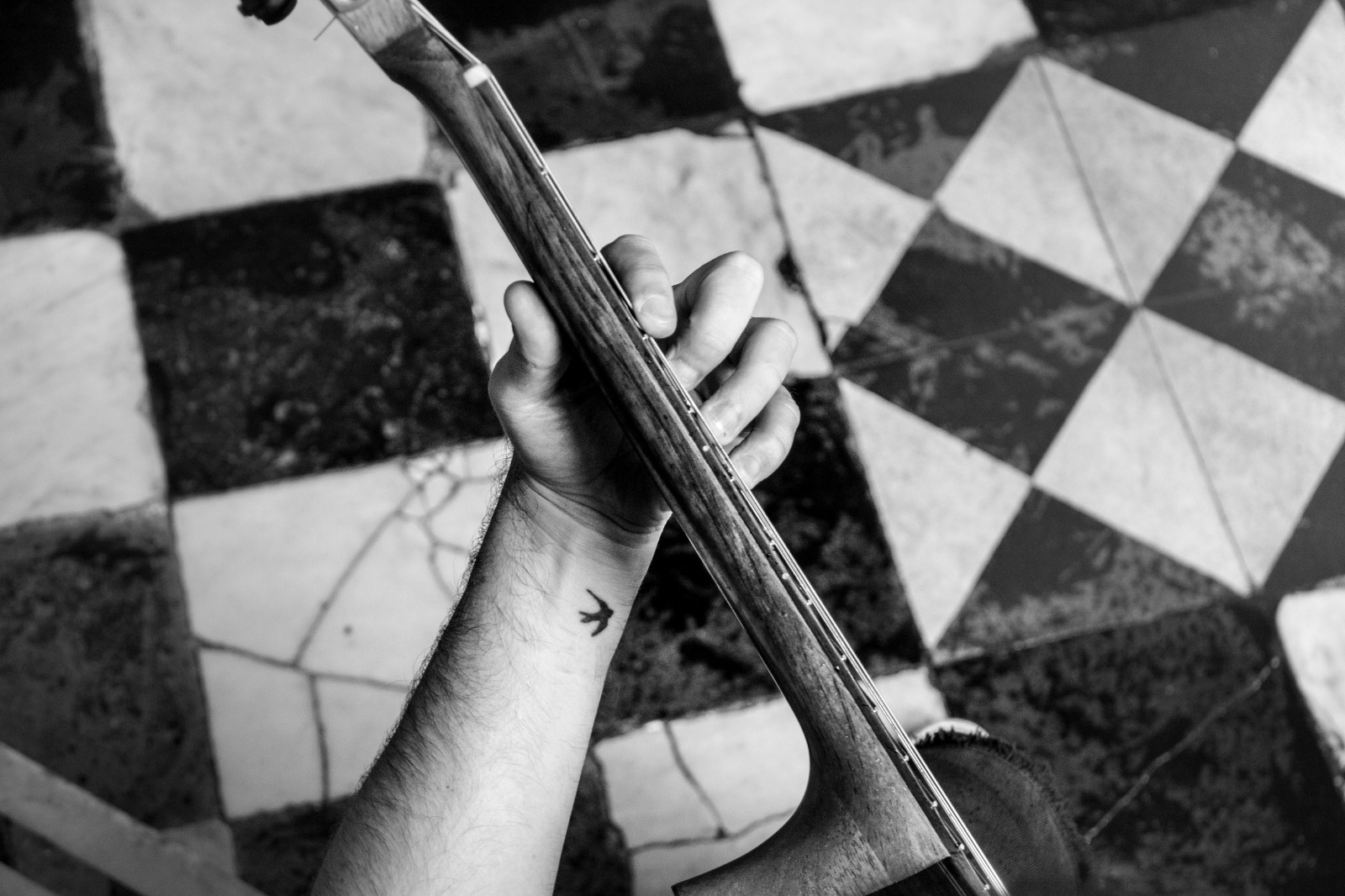 A hand on the neck of a guitar above a cracked checkered tile floor