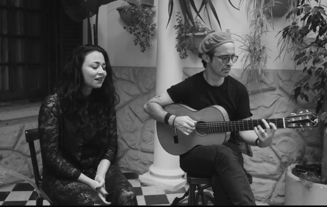 A singer and a guitarist seated side by side in a courtyard with checkered tiles